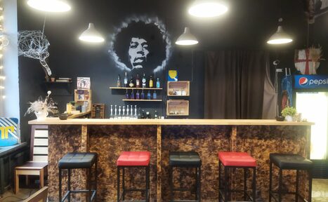 Арендовать Лофт-Бар. Барная стойка с пятью стульями, чередующимися чёрными и красными сиденьями. На темной стене висит изображение человека. Справа холодильник для напитков. Освещение представлено подвесными светильниками. На стойке и полках алкогольные бутылки.