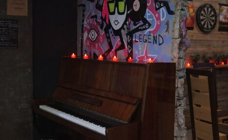 Арендовать Лофт-Бар. Кафе или бар с угловым расположением настенного пианино, рядом рисунок граффити на стене. На пианино стоят небольшие свечи. На стене справа дартс и деревянная обшивка. Оттенки коричневого, красного, фиолетового и синего.