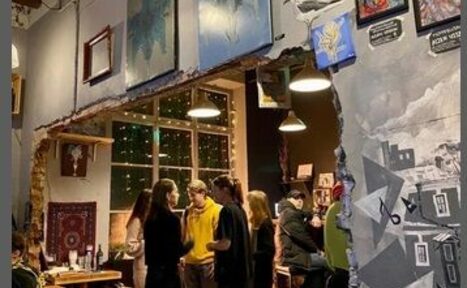 Арендовать Лофт-Бар. Творческое пространство с арт-декором, картинами на стенах, открытая кирпичная кладка, подвесные светильники, винные бутылки на столах, люди общаются, деревянная мебель, большая декорированная стена с рисунками, большое окно с гирляндами за стеклом.