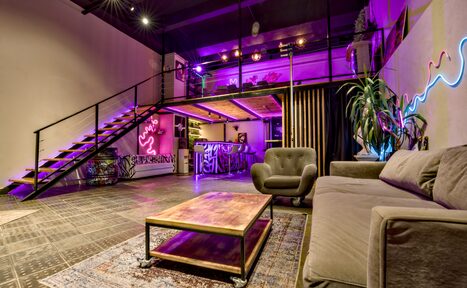 Арендовать НЕОН Лофт. Лофт-пространство с мезонином, металлической лестницей, фиолетовой подсветкой. На первом уровне серый диван, кресло, деревянный стол. На втором уровне барная зона. Зелёные растения и неоновая надпись на стене. Тёмный потолок и пол.