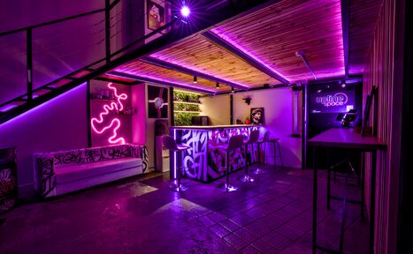 Арендовать НЕОН Лофт. Барное пространство с неоновым освещением, лиловыми акцентами. Барная стойка украшена граффити, несколько высоких стульев. Слева белый диван, на стене неоновый знак, пол бетонный, потолок деревянный. Видна лестница на второй уровень.