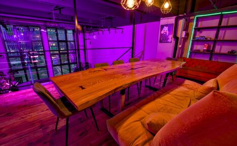 Арендовать НЕОН Лофт. Просторная комната с длинным деревянным столом и стульями, красный диван, полки на стене. Фиолетовое и зеленое освещение, открытая планировка. На столе стоят светильники с открытыми лампами. Деревянный пол, крупные окна.