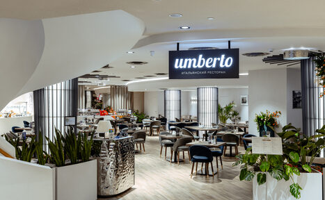 Арендовать Итальянский ресторан Umberto(Умберто). Современный ресторан с открытой планировкой, круглые столы и мягкие стулья, растения в белых горшках, светлые деревянные полы, круглые потолочные светильники, металлическая барная стойка, черный подвесной знак с надписью "умберто итальянский ресторан".