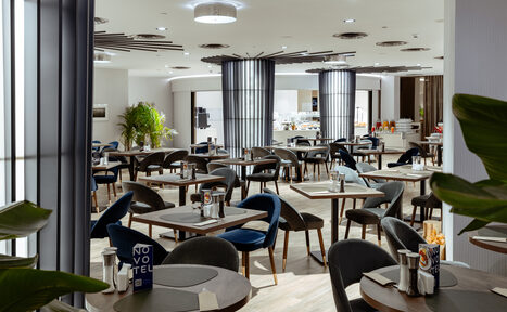 Арендовать Итальянский ресторан Umberto(Умберто). Современный ресторан с круглыми и квадратными столами, темно-синими стульями, мягким освещением. Светлая отделка, деревянные полы, декоративные растения. Пространство для фуршета с напитками и закусками на фоне, надпись "NOVOTEL" на столах.