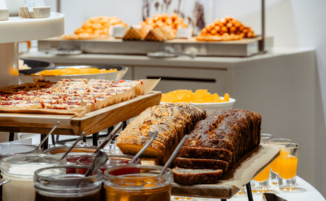 Арендовать Итальянский ресторан Umberto(Умберто). Стол с разнообразными десертами: пироги, кексы и хлеб на подносах, банки с джемом; задний план — апельсиновые шарики и сочные кусочки фруктов. Светлое помещение с нейтральной цветовой палитрой.