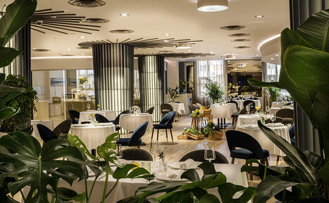 Арендовать Итальянский ресторан Umberto(Умберто). Пространство ресторана с круглым столами, накрытыми белыми скатертями, темными стульями, зелеными растениями. Светлые стены, подвесные потолочные светильники, деревянный пол. Декор из листьев на первом плане.