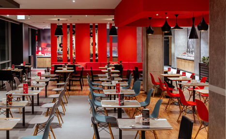 Арендовать Ресторан Ibis Kitchen(Ибис Кухня). Ресторан с открытой планировкой, множество столов и стульев; черные и красные кресла, белые столы. Яркое освещение, красные и серые стены, деревянный пол. Черные подвесные светильники, красные акценты, белый потолок.