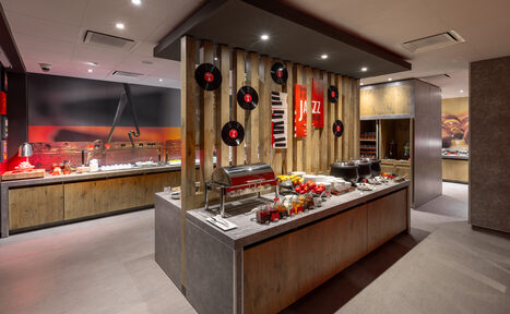 Арендовать Ресторан Ibis Kitchen(Ибис Кухня). Кейтеринговое помещение с деревянными панелями и серыми столешницами. На стене виниловые пластинки и плакат с надписью "Jazz". На столе еда и посуда. Потолочные светильники, светло-серое напольное покрытие.