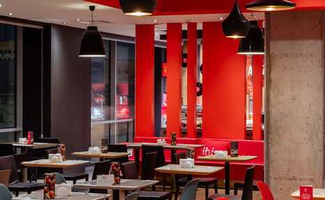 Арендовать Ресторан Ibis Kitchen(Ибис Кухня). Современное кафе с красными и черными акцентами. Столы с черными и серыми стульями, большие окна. Потолочные светильники, бетонная стена, декоративные панели, меню на столах. Яркое освещение, чистое состояние.