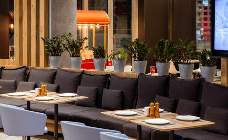 Арендовать Ресторан Ibis Kitchen(Ибис Кухня). Помещение ресторана с длинным диваном, деревянными столами и белыми стульями. На столах белая посуда и перечницы. Сзади дивана растения в серых горшках. На стене телевизор. Яркое освещение. В углу красный попкорн-аппарат.