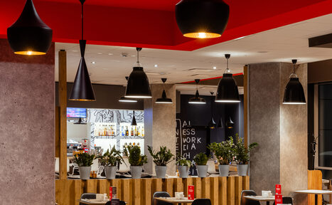 Арендовать Ресторан Ibis Kitchen(Ибис Кухня). Кафе с деревянными столами и стульями, оформленными в серых и черных тонах. На столах меню в красных держателях. Потолок белый и красный, светильники черные подвесные. Присутствуют растения в белых горшках. На стене текстовое граффити.