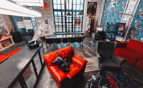 Арендовать Лофт Делюкс. Лофт-пространство с крупными окнами, деревянный пол, длинный стол с черными стульями, красное и серое кресла, мягкая игрушка, большая люстра, открытый гардероб с белыми платьями, телевизор, голубые обои с узором.