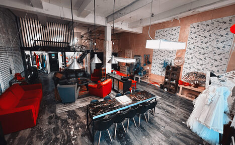 Арендовать Лофт Делюкс. Лофт-пространство с высокими потолками и белыми балками. Красный и серый диваны, длинный стол с черными стульями, промышленное освещение. Стена из кирпича, декор из растений и картин. Гардеробная зона и большое зеркало. Темный деревянный пол.