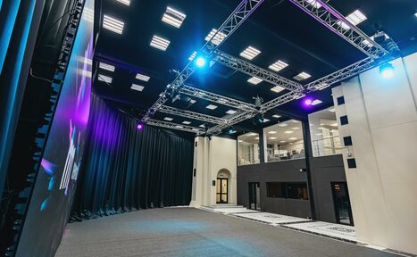 Арендовать New Media (Нью Медиа) продакшн. Павильон с большим LED-экраном. Просторное помещение с высокими потолками и сценой. На стене большой экран. По периметру установлены световые конструкции с прожекторами. Пол ковровый, стены светлые с черными акцентами. Сбоку два уровня с окнами и дверями.