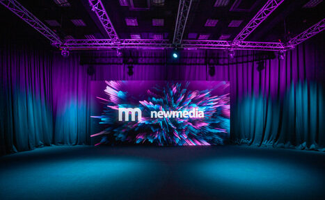 Арендовать New Media (Нью Медиа) продакшн. Павильон с большим LED-экраном. Просторное помещение с черными тканевыми драпировками по периметру и сценой. В центре яркий экран с абстрактным дизайном и текстом "newmedia". Потолок с подвесной конструкцией и фиолетовым освещением. Пол темного цвета.