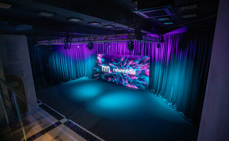 Арендовать New Media (Нью Медиа) продакшн. Павильон с большим LED-экраном. Сценическое пространство с темными шторами и экраном, на котором виден текст "newmedia". Освещение фиолетового и синего оттенков, потолок с квадратными светильниками. Помещение в хорошем состоянии.