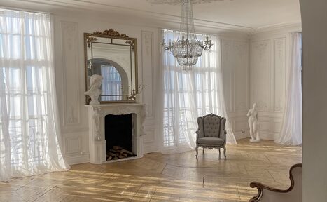 Арендовать Арт-Плейс Фактура. Просторный лофт в классическом стиле. Светлая комната с высокими потолками, паркетным полом. У стены декоративный камин с зеркалом и бюстом. На потолке люстра. У окна серое кресло. Боковые стены украшены лепниной, имеются плинтусы. Шторы из легкой ткани.