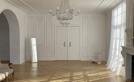 Арендовать Арт-Плейс Фактура. Просторный лофт в классическом стиле. Просторная светлая комната с белыми стенами и высокими потолками. На полу паркет, крупная люстра, арка, белые двустворчатые двери. У стены стоит зеркало, у окна бюст, рядом белая колонка. Длинные полупрозрачные шторы закрывают большое окно.