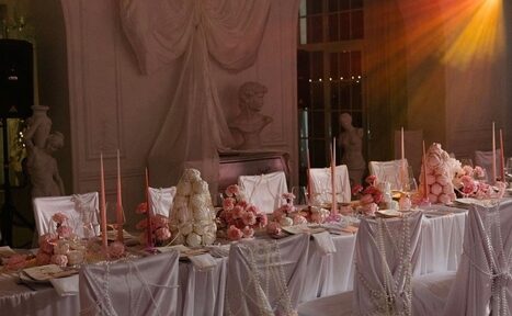 Арендовать Арт-Плейс Фактура. Просторный лофт в классическом стиле. Банкетный зал с длинным накрытым столом, белые скатерти и чехлы на стульях, декор из розовых цветов и свечей, скульптуры на стенах, мягкое освещение теплых тонов, на стене белый архитектурный декор и бюст.