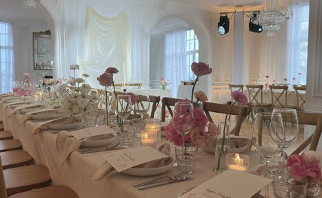 Арендовать Арт-Плейс Фактура. Просторный лофт в классическом стиле. Просторная банкетная комната с длинным столом, накрытым светлой скатертью, украшенным розовыми и белыми цветами. Деревянные стулья, свечи, бокалы, карточки на тарелках. Белые стены, арочные окна с белыми шторами, хрустальная люстра на потолке, светлое напольное покрытие.
