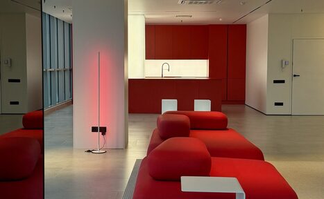 Арендовать Арт-Плейс Фактура. Современный лофт с видом на весь город. Современное пространство с открытой планировкой, красной кухней и деревянным полом. Красные диваны с журнальным столиком, высокие окна, вертикальное освещение. Светлые стены, потолочные светильники, дверь сбоку, минималистичный дизайн.