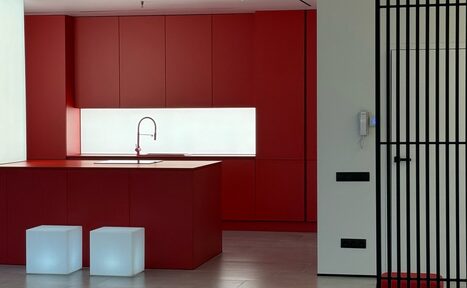 Арендовать Арт-Плейс Фактура. Современный лофт с видом на весь город. Кухонное пространство с красными шкафами и островом, встроенная мойка с изливом, светлое фоновое освещение, два белых кубических сиденья, светлый деревянный пол, черные решётки перегородки и настенный домофон.