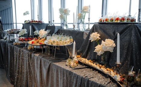 Арендовать Арт-Плейс Фактура. Современный лофт с видом на весь город. Стол с серой блестящей скатертью, на котором расставлены вазы с белыми цветами, высокие белые свечи и блюда с канапе и десертами. Стены с крупными окнами пропускающими много света.