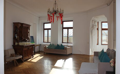 Арендовать Cassiopea (Кассиопея). Светлая гостиная с деревянным паркетом, два дивана с подушками, антикварный шкаф, стулья. Большие окна, люстра с красными элементами. Белые стены, скульптурная ниша. Мягкий дневной свет.