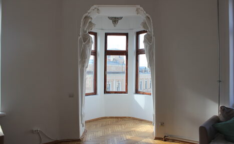 Арендовать Cassiopea (Кассиопея). Зона в светлой комнате с паркетом, декором в виде скульптур на арке; эркер с тремя окнами, вид на городские здания; справа часть дивана.