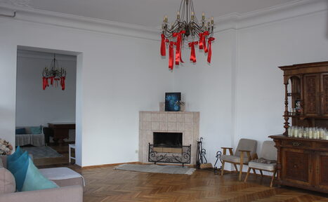 Арендовать Cassiopea (Кассиопея). Гостиная с паркетным полом и белыми стенами. В центре камин с декоративной решёткой, над ним картина. Справа деревянный буфет. Диван с цветными подушками, два кресла и столик. Две люстры с красными лентами. Освещение естественное.