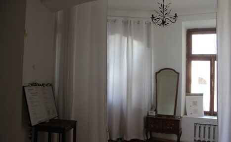 Арендовать Cassiopea (Кассиопея). Комната с деревянным полом, белыми занавесками, потолочным светильником, старинным зеркалом на комоде. На стене окно с деревянной рамой и радиатором. Справа от зеркала настольный мольберт с картиной. Слева столик с меню.