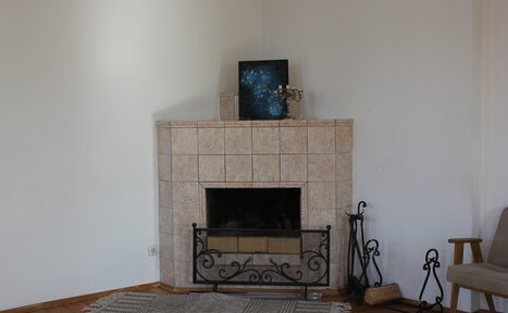 Арендовать Cassiopea (Кассиопея). Комната с камином, облицованным светлой плиткой. Вокруг него декоративная кованая решетка, рядом инструменты для камина. На камине картина и подсвечник. На полу светлый ковер, рядом кресло. Стены белые.