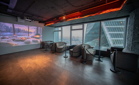 Арендовать ATMOSFERA 53(Атмосфера 53). Просторное помещение с окнами на всю высоту стены, с видом на город. Справа стоят круглые кресла и прямоугольные столики, разделенные барьерами. На стене слева проектируется изображение зимнего пейзажа. Подвесное освещение, темный пол, бетонный потолок.