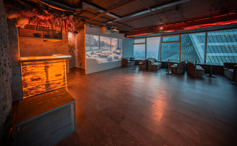 Арендовать ATMOSFERA 53(Атмосфера 53). Просторное помещение с большими окнами и видом на город. Слева – барная стойка с подсветкой. У стены – проектор с изображением зимнего пейзажа. Справа несколько кресел и столиков. Потолок с открытыми коммуникациями. Освещение тёплое, рассеянное.