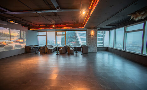 Арендовать ATMOSFERA 53(Атмосфера 53). Просторное помещение с панорамными окнами и видом на город. Темный пол, серые стены, потолок с декоративной подсветкой. Несколько мягких кресел и столов у окна. На стене проекция изображения горящего камина.