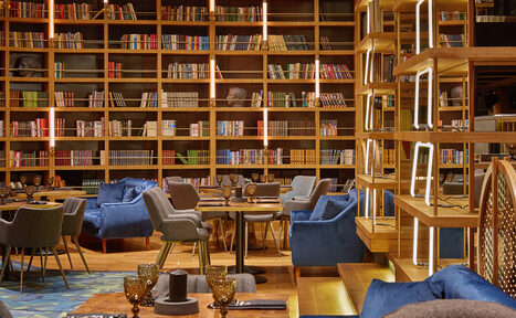 Арендовать Communitymoscow (Коммьюнити Москоу).Зал Библиотека. Многофкнкциональное пространство. Библиотечное пространство с высокой деревянной полкой, заполненной книгами. В центре расставлены круглые столы и серо-синие кресла. Потолок и пол оформлены деревянными элементами. Освещение мягкое, с акцентными светильниками.