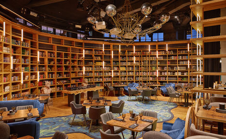 Арендовать Communitymoscow (Коммьюнити Москоу).Зал Библиотека. Многофкнкциональное пространство. Библиотечное пространство с деревянными стеллажами, заполненными книгами. Планировка с креслами и столами для отдыха и работы. Темный потолок с современными светильниками. Пол с узорчатым ковром. Приятное теплое освещение, на стенах лампы.