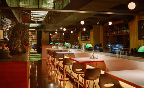 Арендовать Communitymoscow (Коммьюнити Москоу).Зал Библиотека. Многофкнкциональное пространство. Барное пространство с длинной стойкой и высокими стульями, полки с напитками на стене. Подвесные круглые светильники, настольные лампы с зелеными абажурами. Темное деревянное и металлическое оформление, зеркальные потолки и светящаяся надпись.