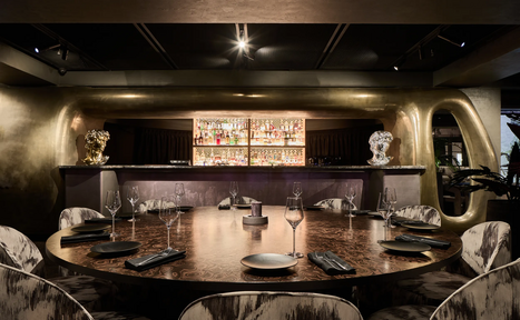 Арендовать Communitymoscow (Коммьюнити Москоу).Зал Библиотека. Многофкнкциональное пространство. Круглый стол с несколькими стульями, сервированный пустыми тарелками и бокалами. На заднем плане барная стойка с полками для бутылок. Потолочное освещение, стены и бар отделаны в золотистых тонах. Темное общее освещение, современный интерьер.