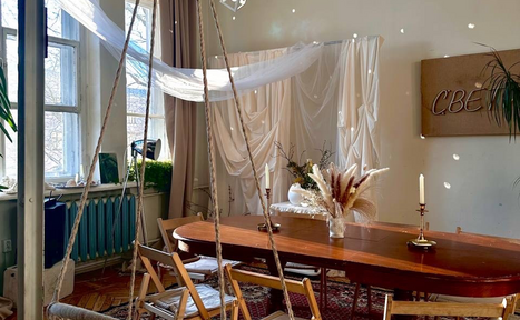 Арендовать Студия Свети. Пространство с деревянным столом, окружённым складными стульями. На столе свечи и ваза с сухоцветами. Светлая ткань подвешена у окна и на стене. Окно с занавесками, синий радиатор. На стене знак "СВЕТ". Паркетный пол, ковёр.