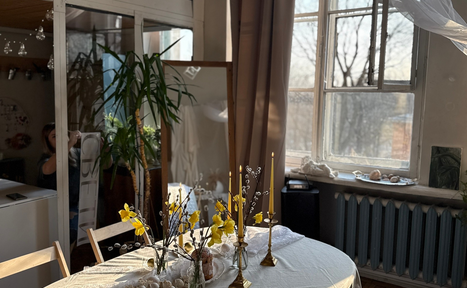 Арендовать Студия Свети. Просторная комната с круглым столом, покрытым белой скатертью, в центре цветочные декорации и золотые подсвечники. Возле окна с бежевыми шторами радиатор отопления. Зеркало и крупное растение в углу. Светлый и уютный интерьер.