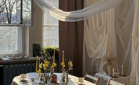 Арендовать Студия Свети. Просторная комната с большими окнами и открытыми занавесками. В центре стола белая скатерть, на которой расположены свечи, цветы и кофейные чашки. На фоне драпировки из белой ткани. Естественное освещение, дерево и текстиль в декоре.