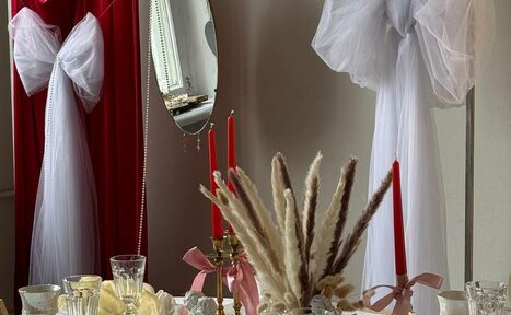 Арендовать Студия Свети. Столовая с круглым столом, на котором расположены украшения из сухоцветов, свечи в подсвечниках и стеклянная посуда. На стенах красные и белые занавеси. За столом зеркало на стене.