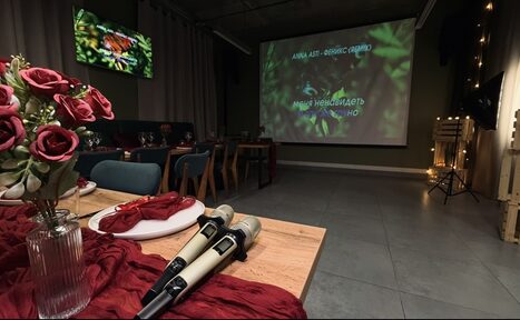 Арендовать Лофт Интра. Зал Фрей. Просторное помещение с проектором, показывающим текст песни. Есть стол с красными украшениями, букетом роз и двумя микрофонами. На стене за столом висит второй экран. Стулья мягкие, темно-синие. Пол облицован плиткой. Освещение приглушенное.