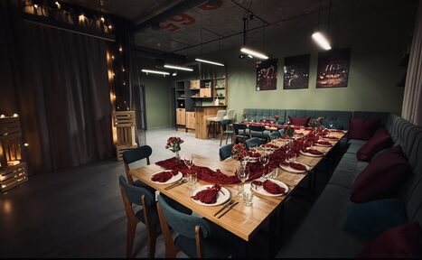 Арендовать Лофт Интра. Зал Фрей. Современное пространство с длинным деревянным столом, сервированным на десять персон, стол украшен красными салфетками и цветами. Слева занавеска, мягкий угловой диван вдоль стен, столы и деревянные стулья, открытые стеллажи. Освещение центральное и подвесное.