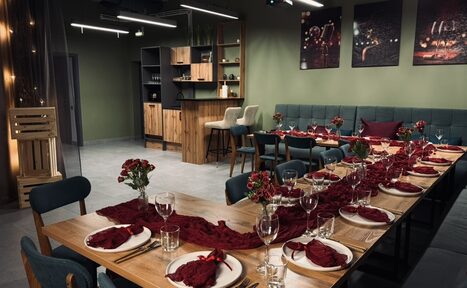 Арендовать Лофт Интра. Зал Фрей. Современное пространство с длинным столом, сервированным на 18 персон. Декорировано тканью и цветами бордового цвета. Барная стойка с высокими стульями, мягкие диваны зелёного цвета. На стене зелёного цвета висят картины. Потолочное освещение.