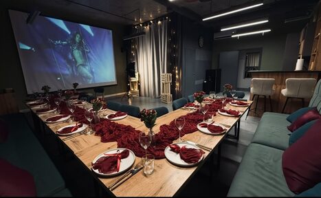 Арендовать Лофт Интра. Зал Фрей. Просторная комната с U-образным деревянным столом, сервированным красными салфетками и бокалами. На стене напротив - экран с проекцией. Серая штора, мягкие диваны, высокий барный стол, подвесные светильники. Тёмные стены и потолок.