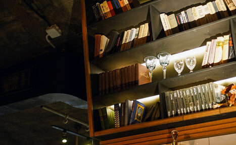 Арендовать Гастробар Дневник. Настенная полка с книгами и бокалами, освещённая тёплым светом. Верхняя часть полки заполнена книгами в разноцветных обложках. На полке также стоят бокалы для вина. Потолок с трубами и софитами.