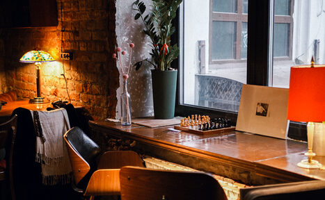 Арендовать Гастробар Дневник. Кофейня с кирпичной стеной, деревянной стойкой у окна, настольными лампами с цветными и красными абажурами. На стойке шахматная доска, декор из растений и сувенирных фигурок. За столом несколько деревянных стульев. Естественное освещение из окна.