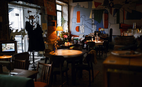 Арендовать Гастробар Дневник. Кафе с круглыми деревянными столами и стульями, настенные росписи, лампа на столе, вешалка с одеждой, за окном видна улица, приглушенное освещение, на столах свечи и бокалы, бетонные стены, тёплые цвета.